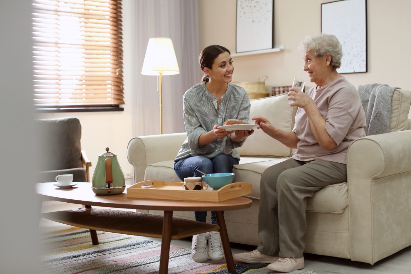 A carer sits with an older woman at home, sharing a drink and conversation.